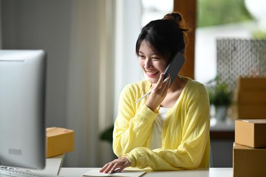 Smiling female small business owner using computer and talking on mobile phone for confirming orders from customer. Online selling, e-commerce concept.