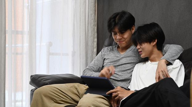 Happy homosexual couple embracing and using digital tablet together on sofa. LGBT and love concept.