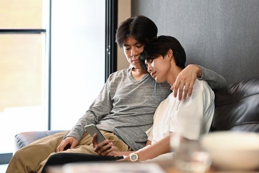 Loving male lgbt couple embracing and using smart phone on sofa together. LGBT and love concept.