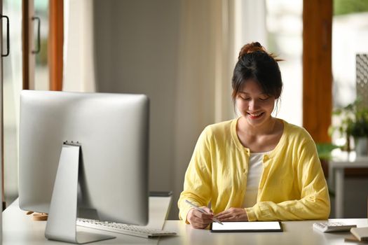 Cheerful asian woman freelancer sitting in bright modern home office and using digital tablet.