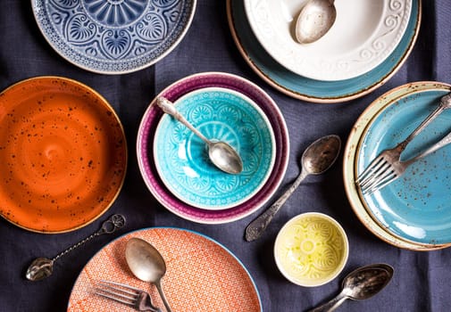 Vintage multicolored empty plates and bowls on a dark gray linen tablecloth. With antique spoons and forks. Table setting. Shabby chic/retro style. Top view. Rustic kitchen