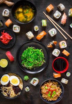 Table served with sushi and traditional japanese food on dark background. Sushi rolls, hiyashi wakame, miso soup, ramen, fried rice with vegetables, nigiri, soy sauce, сhopsticks. Overhead....