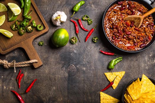 Chili con carne in frying pan on dark wooden background. Ingredients for making Chili con carne. Space for text. Top view. Chili with meat, nachos, lime, hot pepper. Mexican/Texas traditional dish