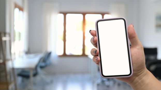 Woman hand holding mock up smart phone with blurred home office background.