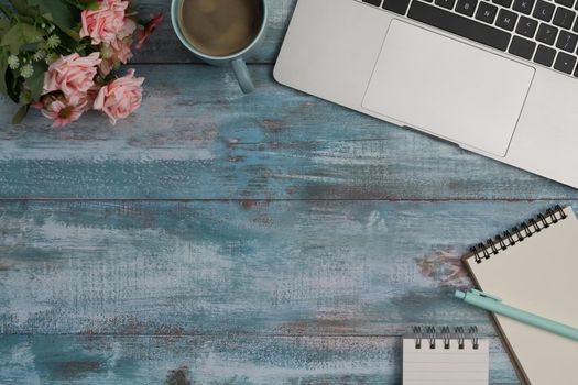 Stylish workplace with laptop computer, notepad, coffee cup and rose flowers on blue wooden desk.