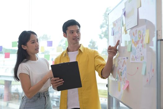 Focused young woman standing near flip chart with her colleague and discussing new marketing strategy together.