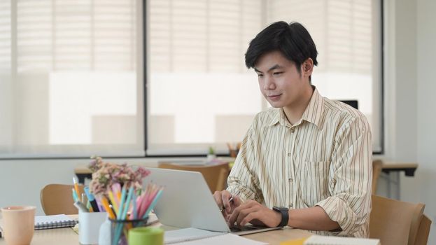 Asian male employee working with computer laptop at modern co working office space.