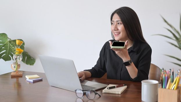 Positive asian woman using laptop computer and having conversation on mobile phone.