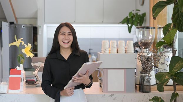 Successful female business owner standing behind counter of coffee shop and smiling to camera.