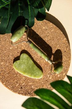 Jade Gua sha scraper and face roller massager on a cork round stand with a monstera leaf. Hard light, shadows, the concept self-care. Facial care. Zero waste. Lifting and toning treatment at home.