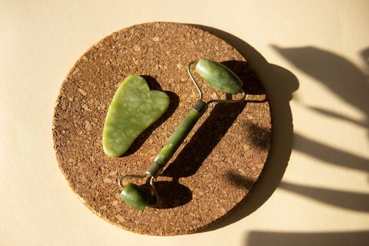 Jade Gua sha scraper and face roller massager on a cork round stand with a monstera leaf. Hard light, shadows, the concept self-care. Facial care. Zero waste. Lifting and toning treatment at home.