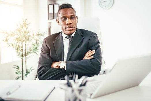 African smiling businessman sitting in the office with crossed arms and looking at laptop.