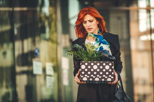 Serious pensive business woman is walking in office district with a box full of her personal belongings from the office just after she got fired. 