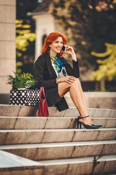 Business woman is sitting on stairs and talking on smartphone next to a box full of her personal belongings from the office just after she gives up job. 