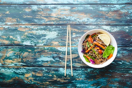 Asian noodles with vegetables, bowl, rustic wooden old background. Space for text. Top view. Soba noodles, vegetables, mushrooms, chopsticks. Vegetarian/Vegan noodles. Asian style dinner with noodles