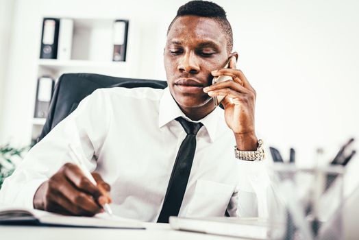 Pensive African businessman is talking on smartphone and writing in notebook in modern office. 