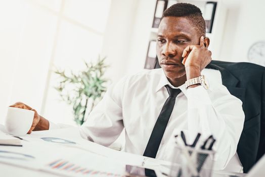 Pensive African businessman working in modern office, drinking coffe, reading  notes and planning what to do next.