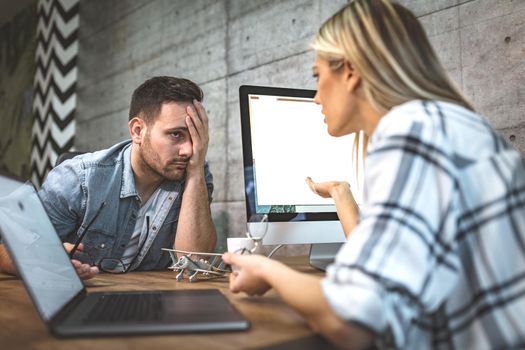 Young handsome frustrated and stressed businessman sitting in the office and complains to his female colleague about the pain in his head.