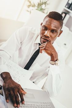 Pensive African businessman is working on laptop in modern office and planning what to do next.