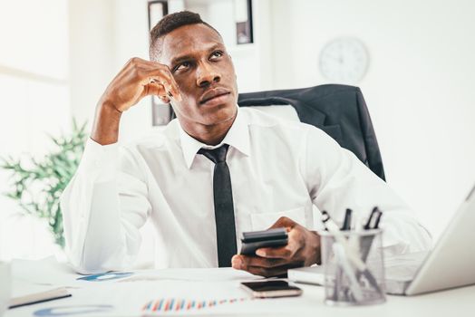 Pensive African businessman working on calculator in modern office and planning what to do next.