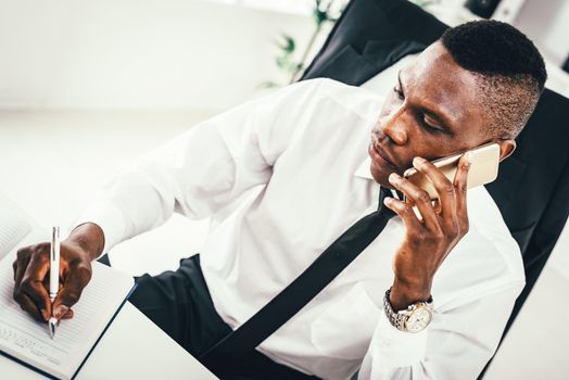 Pensive African businessman is talking on smartphone and writing in notebook in modern office. 