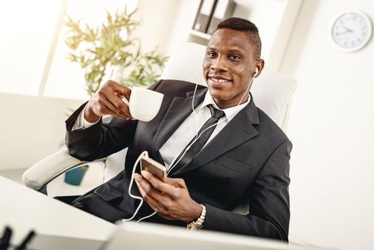 Smiling African businessman is sitting in the office, drinking coffee and listening  music in headphones.