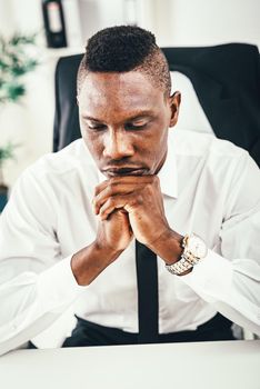 Anxious African businessman sitting in the office and holding his hands under his chin.
