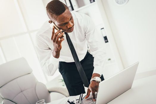 Pensive African businessman talking on smartphone and working at laptop in modern office. 