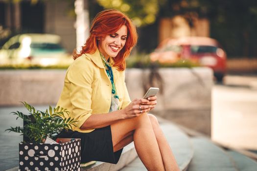 Business woman is sitting on stairs and texting on smartphone next to a box full of her personal belongings from the office just after she gives up job. 