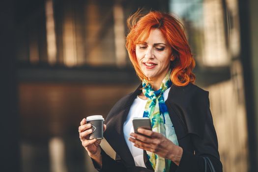 Portrait of a smiling business woman surfing on smartphone on a coffee break in office district at the windy day. She is looking at smartphone with flying hair.