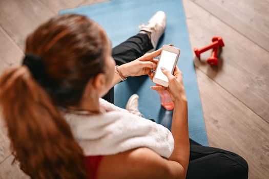 Attractive woman surfing on social media on a smartphone while taking break of exercising in living room at home.