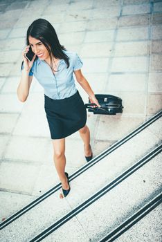 Young beautiful businesswoman standing outdoor with suitcase ready for a business trip and talking on mobile phone.