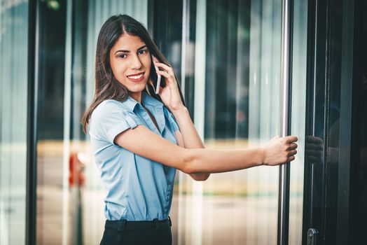 Emotional portrait of a happy and beautiful young businesswoman talking on a smartphone and standing on a business center door.