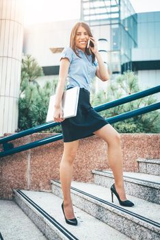 Young beautiful businesswoman walking up the stairs in front of office building, holding a laptop and talking on smartphone.