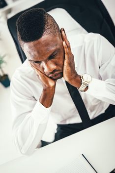 Anxious African businessman sits in the office and holds his head with his hands.