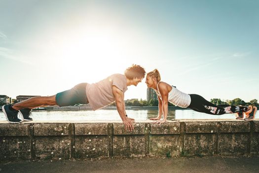 Young happy smiling couple is training outdoors doing push-ups during outdoor cross training by the river.