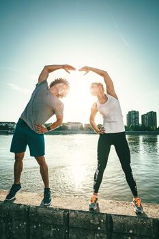 Young happy couple is training outdoors by the river, stretching together on the wall at sunset.