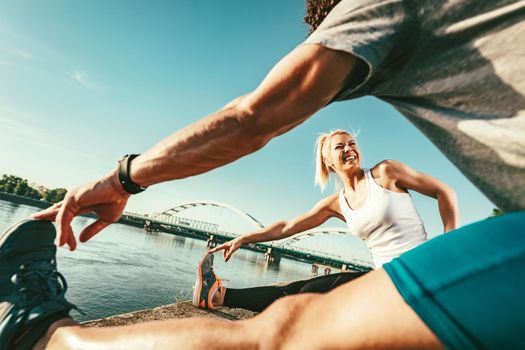 Young happy couple is training outdoors by the river, stretching legs at sunset.