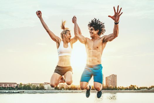 Beautiful young couple is smiling and jumping after successful training by the river in the sunset.