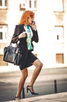 Business woman is walking up stairs to the building with coffee to go. She is talking on smartphone and going to work.