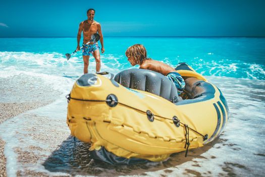 A man is holding a paddle beside a kayak which his son prepares for sailing in the sea.