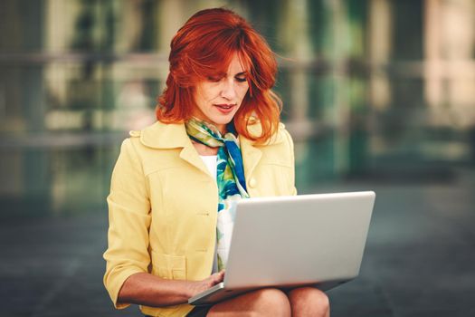 Smiling business woman is sitting on stairs in office district and working at the laptop.