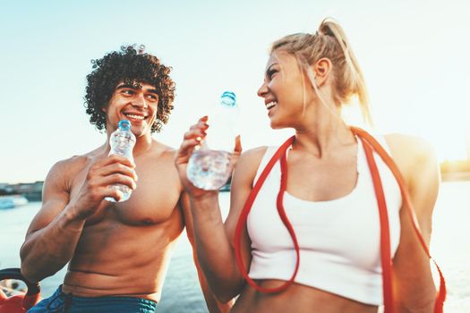 Young fitness couple is resting afther hard training by the river in a sunset drinking water.