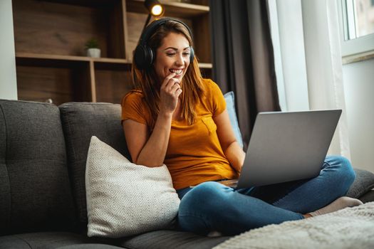 Attractive young woman sitting cross legged on the sofa and using her laptop and headphones to make a video chat with someone at home.