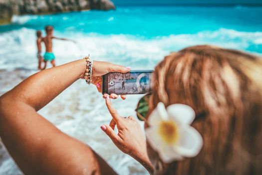 Woman with smartphone takes a photo of her loving sons in sea waves on the tropical beach. Selective focus. Focus on photo on the phone.