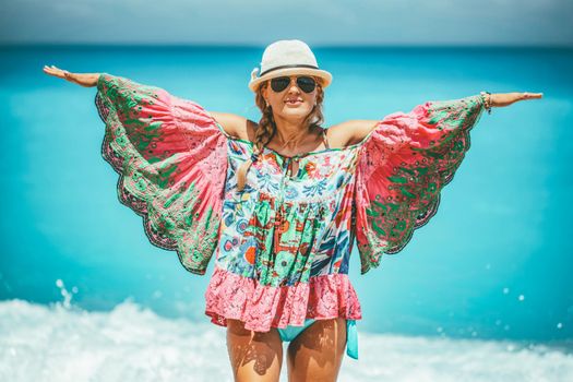 Beautiful young woman in colorful dress with open arms, and summer hat on her head, enjoying at sun in the beach.