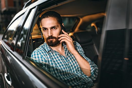 Young confident businessman is holding smartphone in his hand and talking on the back seat in car.