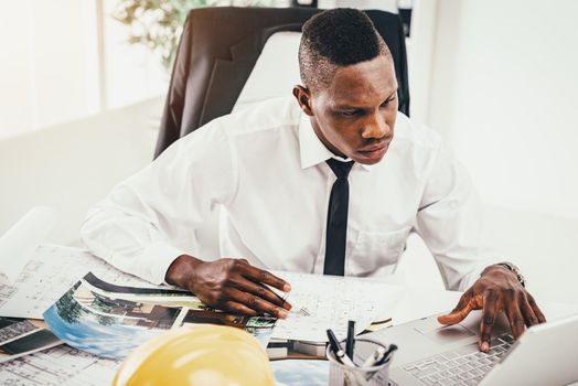 Pensive African architect analyzing blueprint and working on laptop in modern office and planning what to do next.