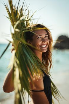 Portrait of an attractive young woman is posing and enjoying at the tropical beach under palm tree leaf.