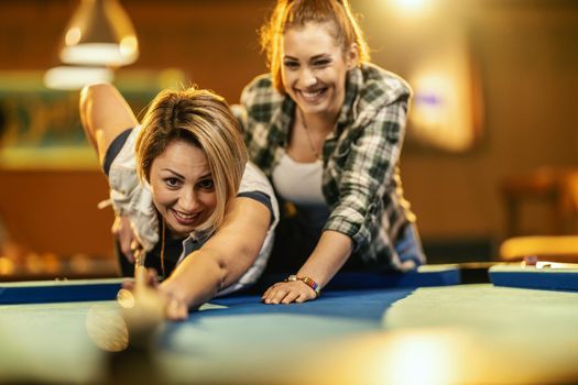 Young smiling cheerful two young women are playing billiards in bar after work. They are involved in recreational activity.